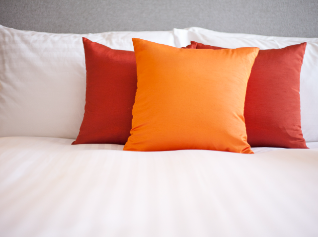 Two red pillows and one orange pillow arranged on a bed with white sheets.